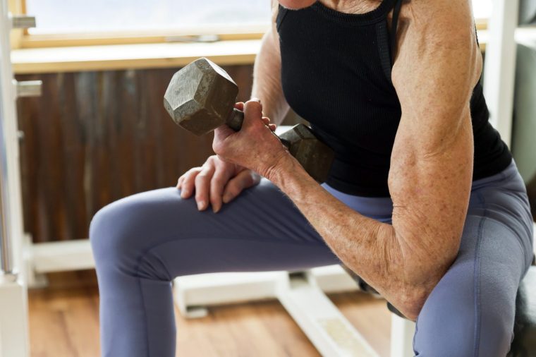 A 76 year old Personal Trainer who has had two prosthetic hips, two prosthetic knees and two shoulder surgeries with an arthritic elbow all caused from her genetics, works out in the weight gym to stay in shape by using a dumbbell to exercise her bicep with curling a dumbbell while sitting on a exercise bench. stock - photo