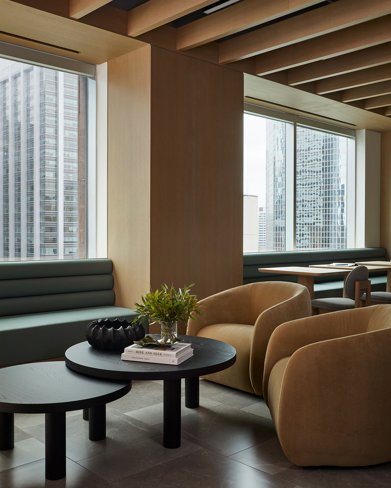Modern office lounge with green cushioned bench seating, tan armchairs, black round tables, books, a plant, and large windows overlooking city skyscrapers.