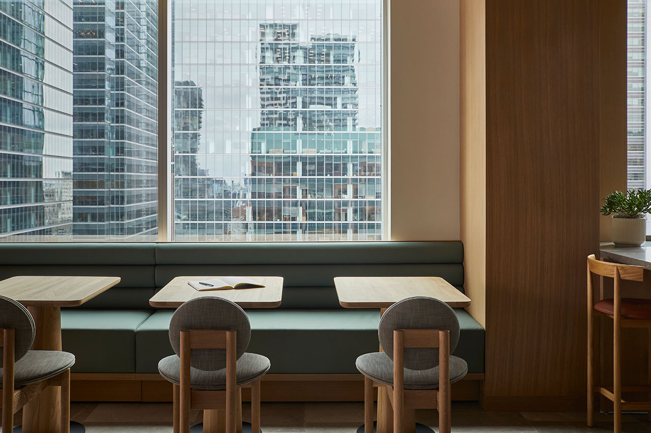 A modern cafe interior with wooden tables and chairs, a long cushioned bench, and large windows showing tall glass office buildings outside.