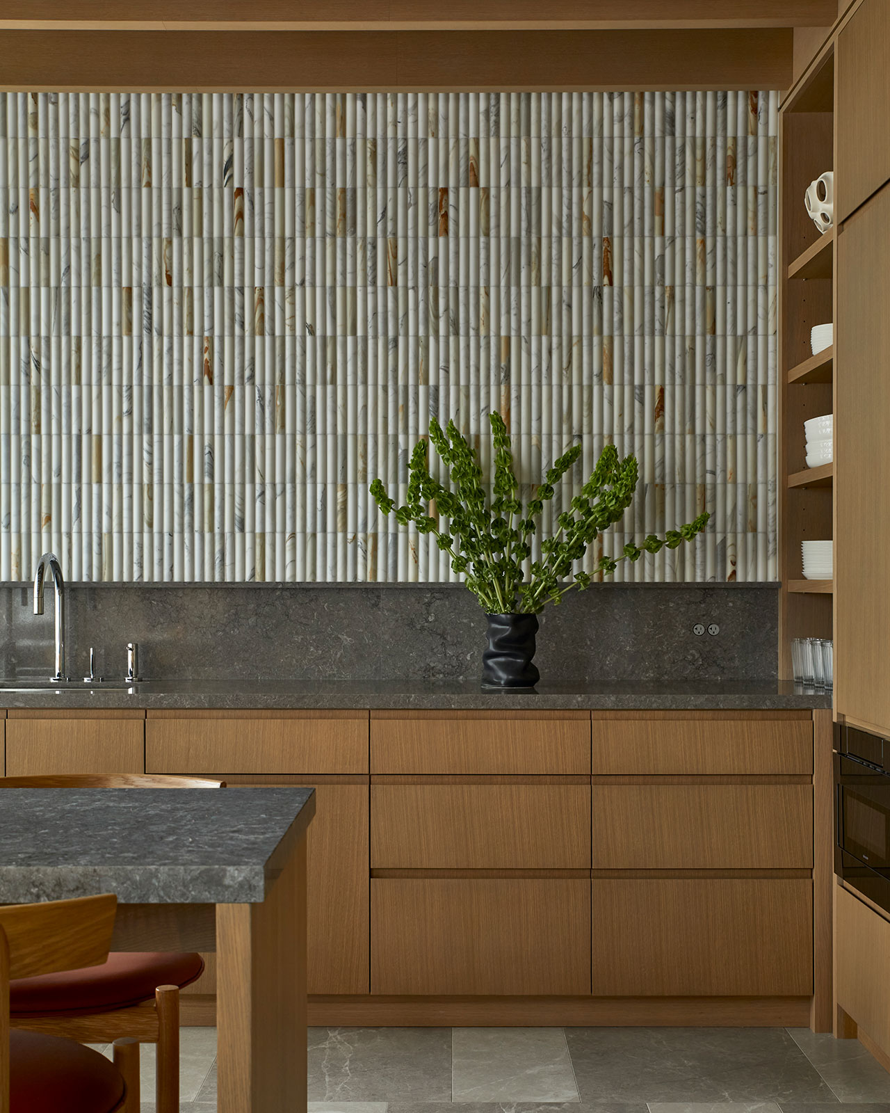 Modern kitchen with light wood cabinets, dark stone countertops, a decorative tiled backsplash, and a leafy green plant in a black vase on the counter.