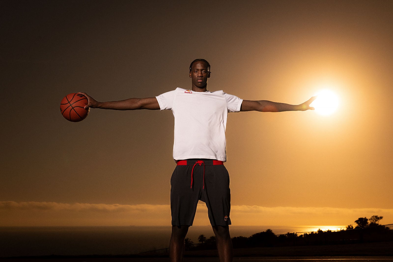 A person stands outdoors at sunset with arms outstretched, holding a basketball in one hand and appearing to hold the Sun in the other, wearing a white shirt and dark shorts.