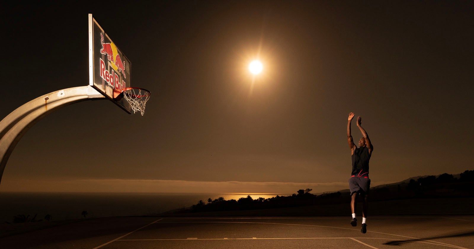 A person jumps to take a basketball shot on an outdoor court at night, with a bright full moon shining in the sky and a Red Bull branded backboard visible. The ocean and silhouettes of trees are in the background.