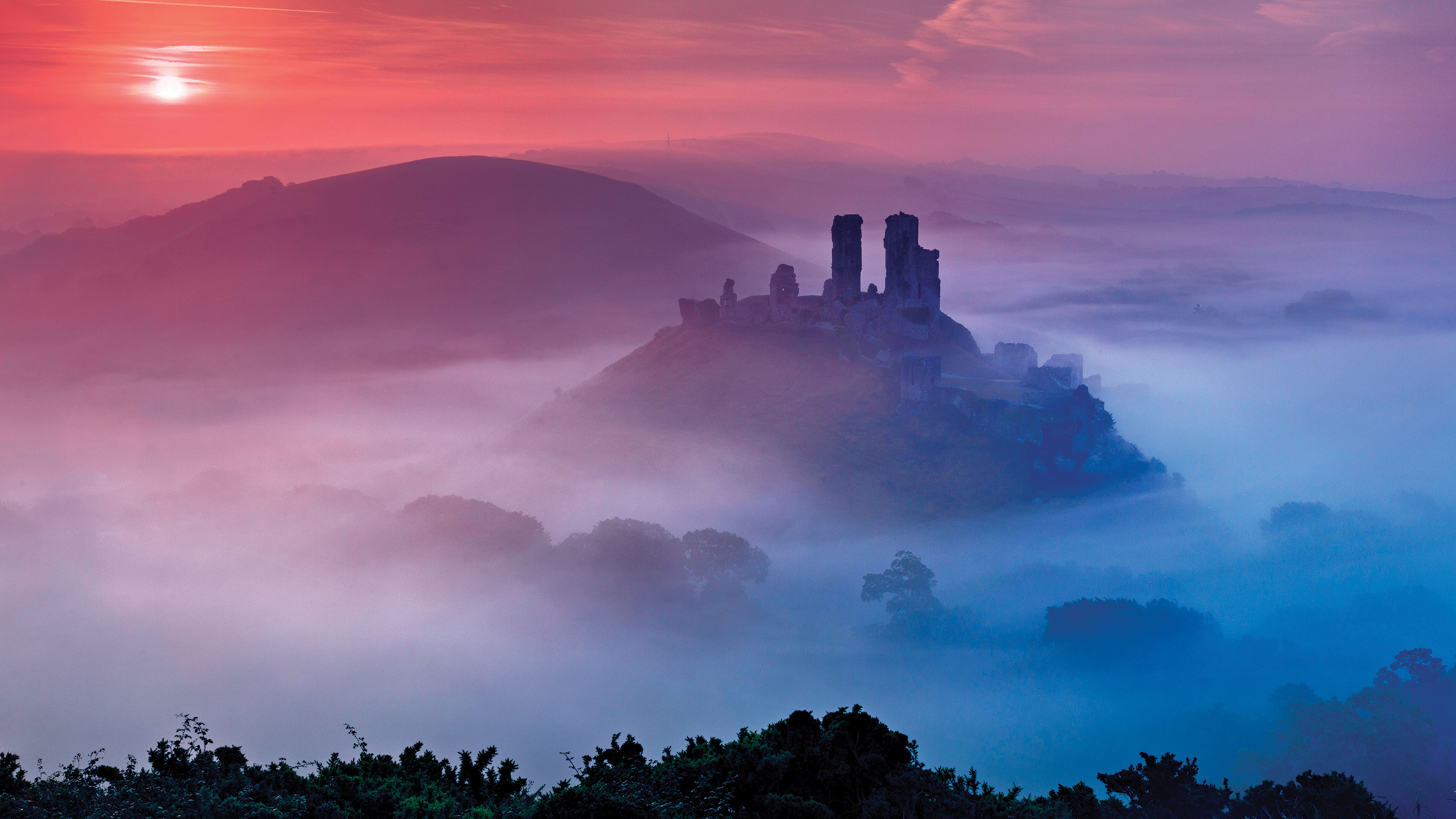 A castle surrounded by fog, which appears purple and pink in the sun, shot from a high perspective