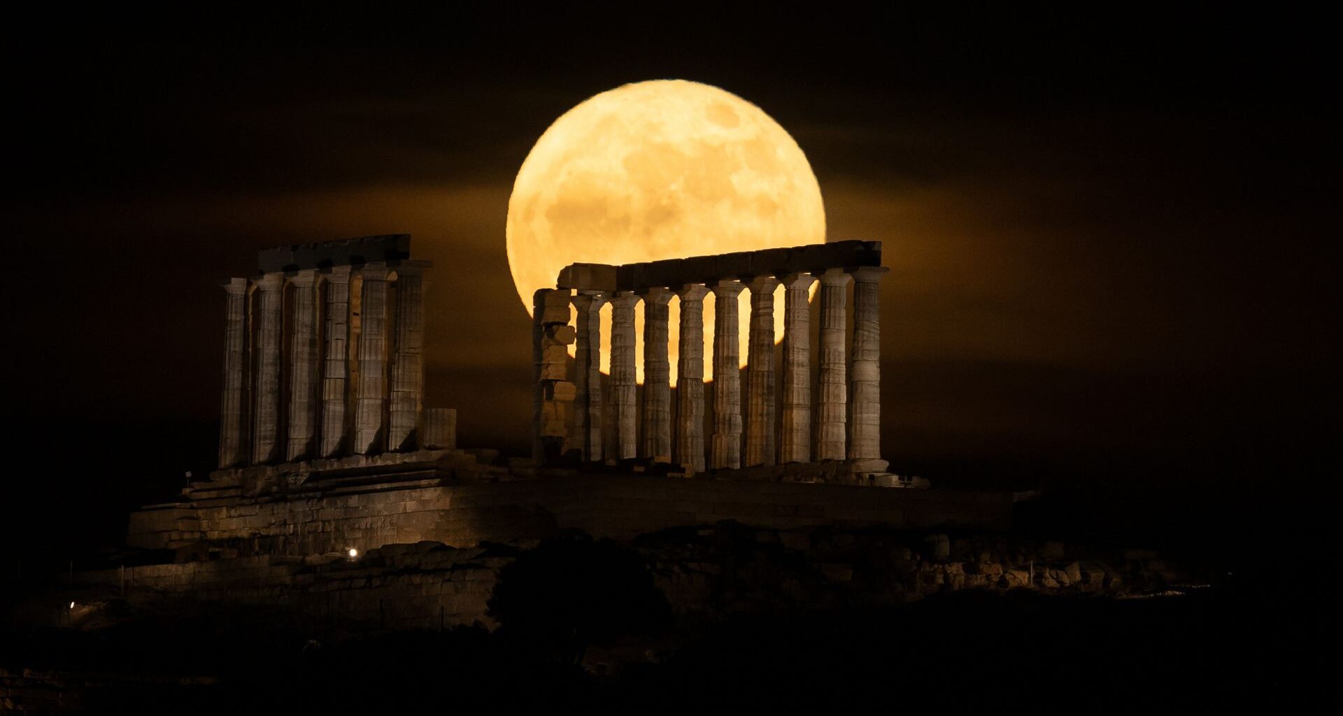 A yellow full moon glows in a dark sky behind the columns of an ancient ruined temple.