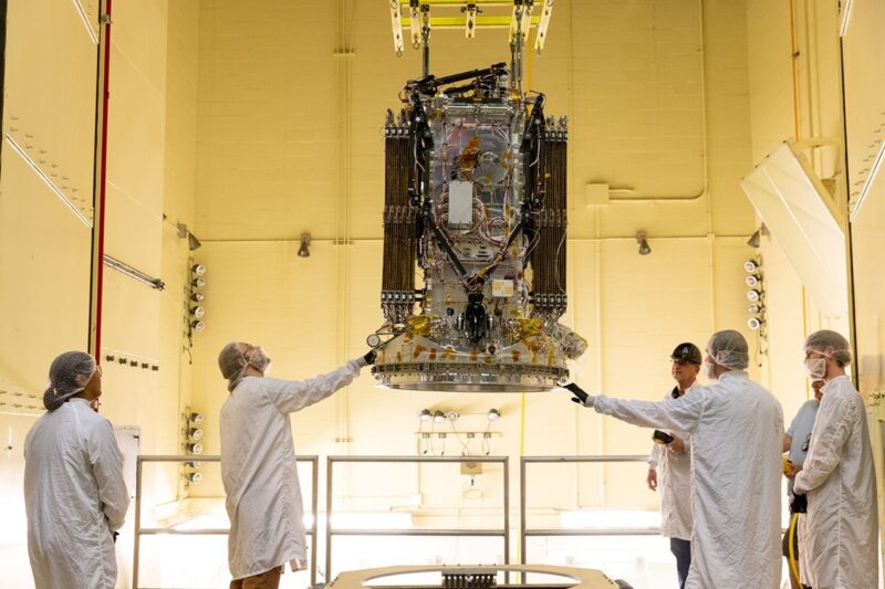 Five people in white suits in a clean room and spacecraft hanging above the floor.