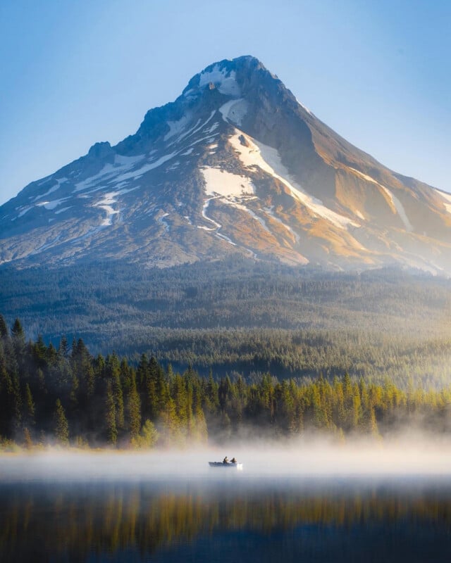 A small boat with two people floats on a misty lake, surrounded by tall evergreen trees, with a snow-capped mountain rising majestically in the background under a clear blue sky.
