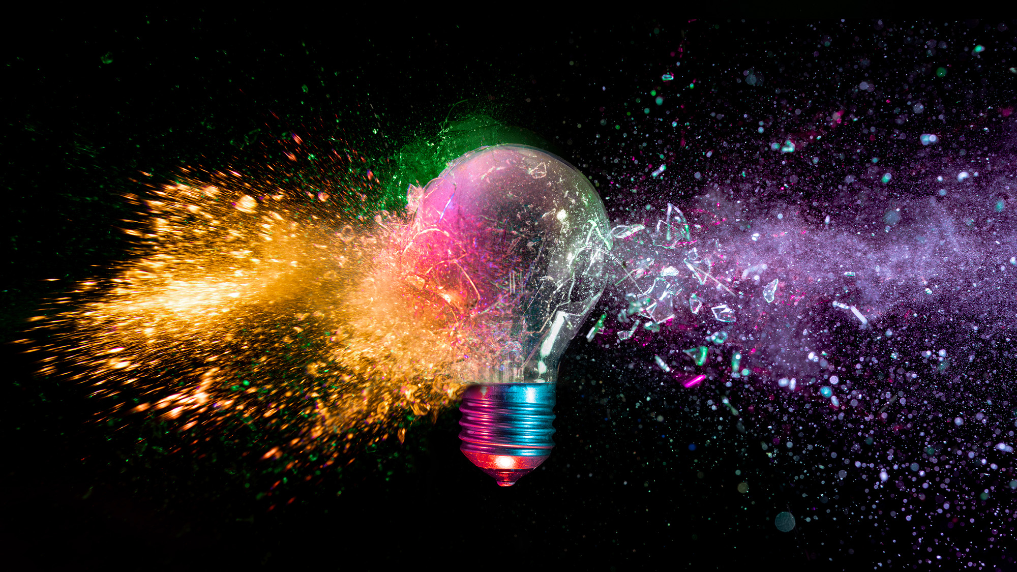 A lightbulb is shattered with various colors coming out of it against a black background