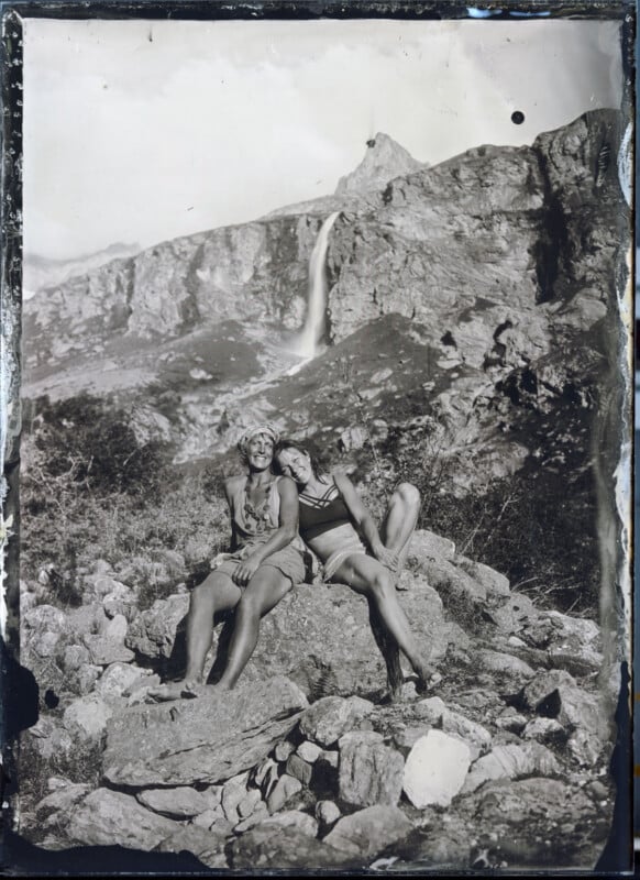 Two people sit on rocky ground, smiling and leaning close together, with a mountain and a waterfall in the background. The image is in black and white and has an old, vintage appearance.