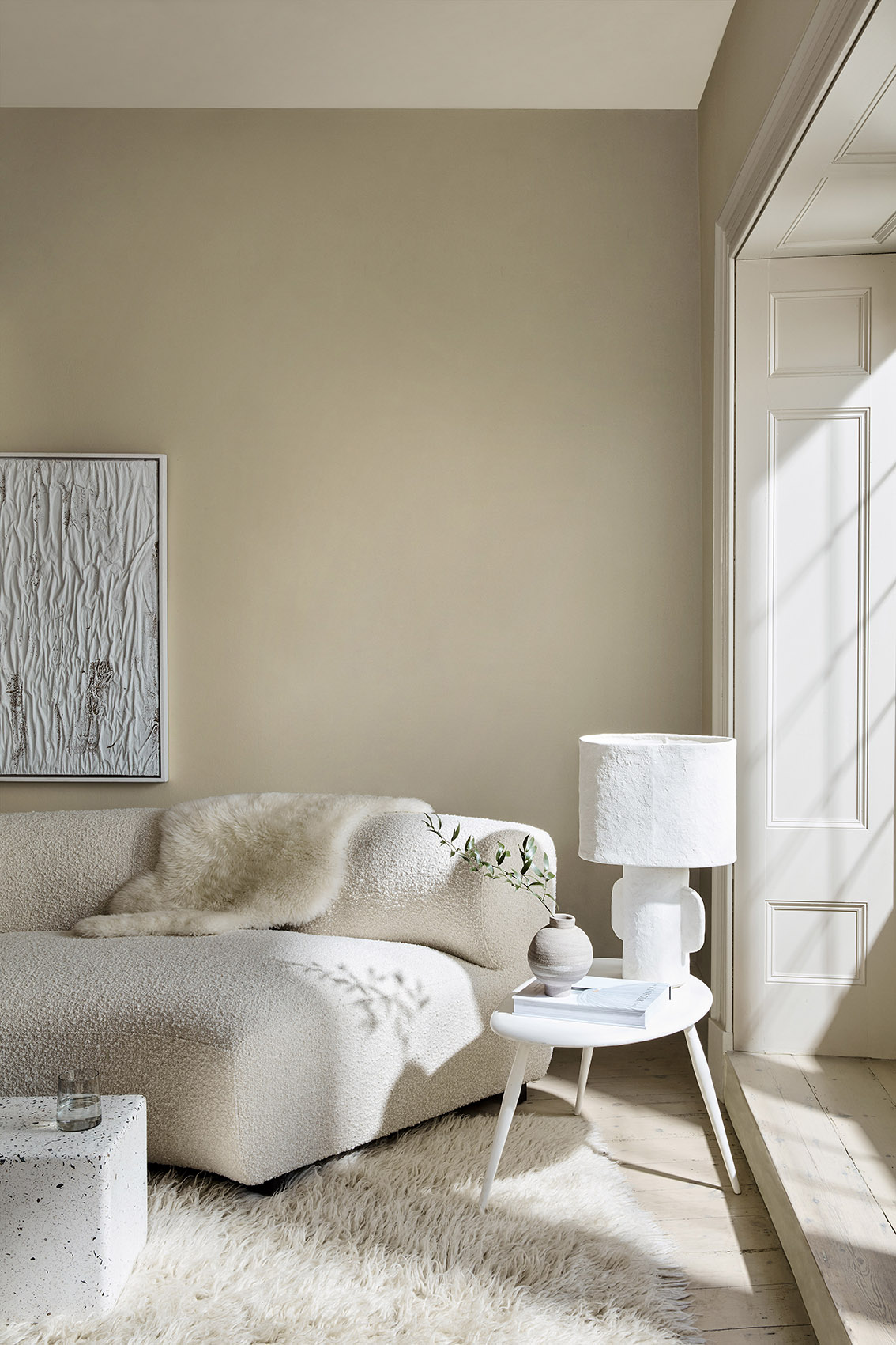 A contemporary living room with sculptural white sofa, white fluffy rug, white side table with white lamp, and cream walls with white textured artwork