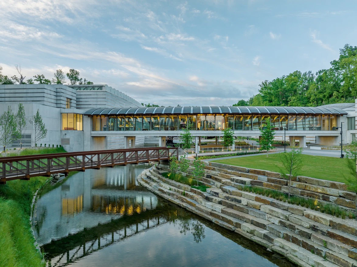 Modern museum with glass facade, surrounded by trees, reflecting on a pond, under a cloudy sky with a wooden footbridge in view.