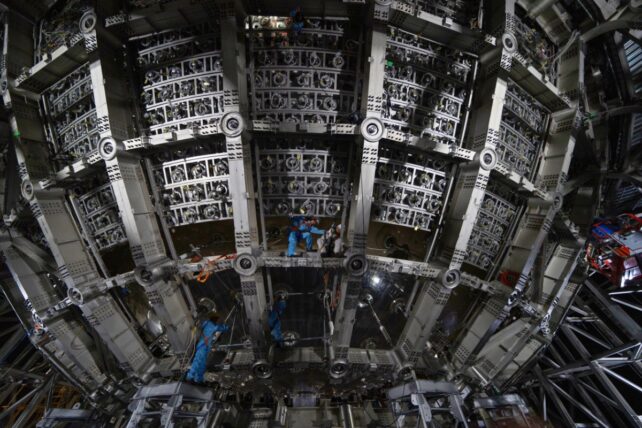 Workers in blue protective suits install components inside the large spherical JUNO detector, surrounded by metal frames and circular sensor mounts.