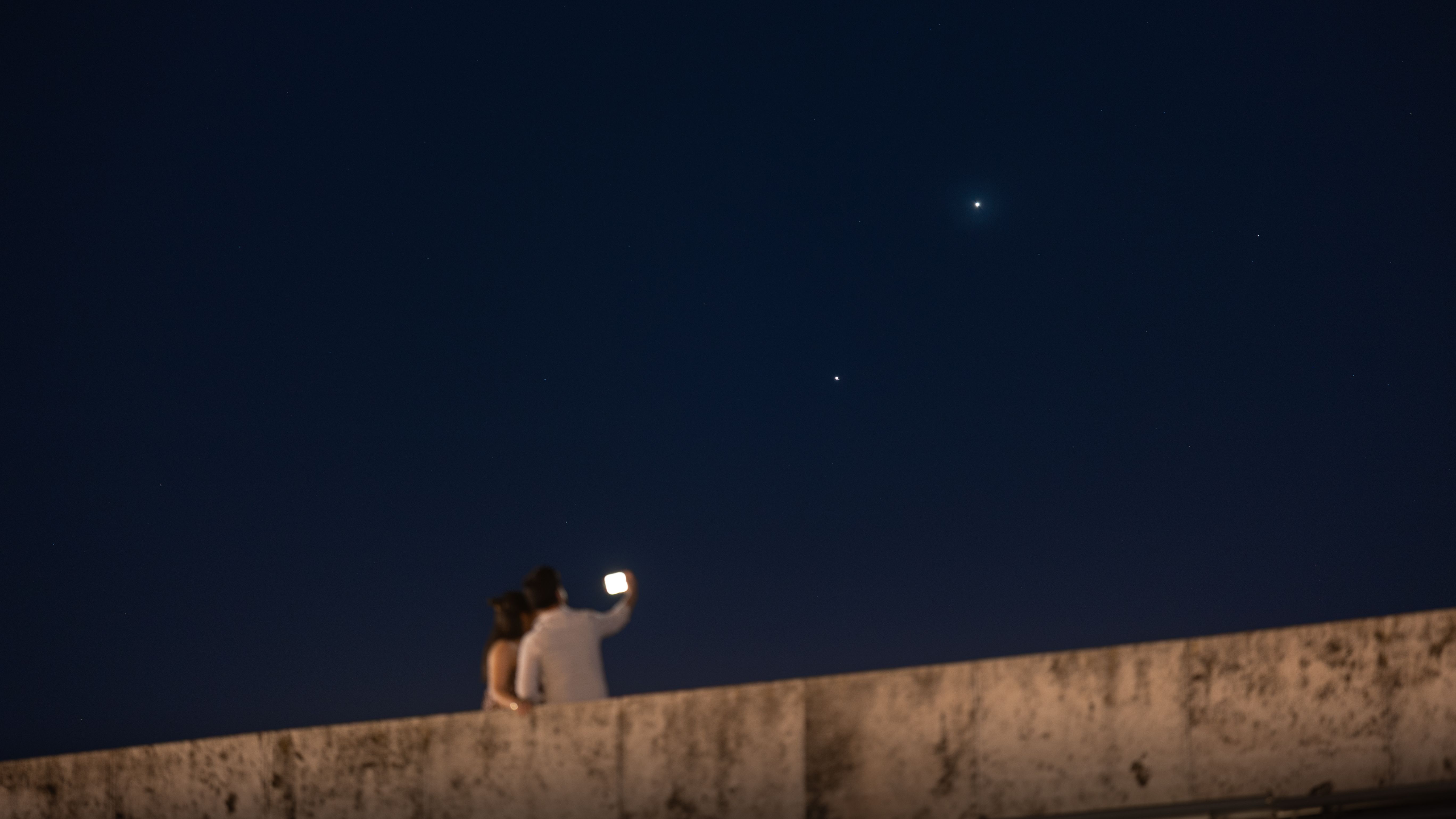 Two star-like planets shine in a dark sky above a man and a woman, who are holding up a phone at the edge of a stone bridge.