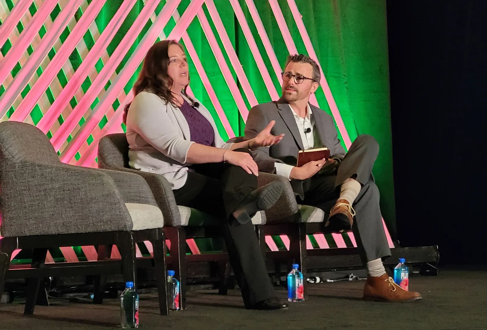 Two people sit in chairs on a stage while addressing an audience.
