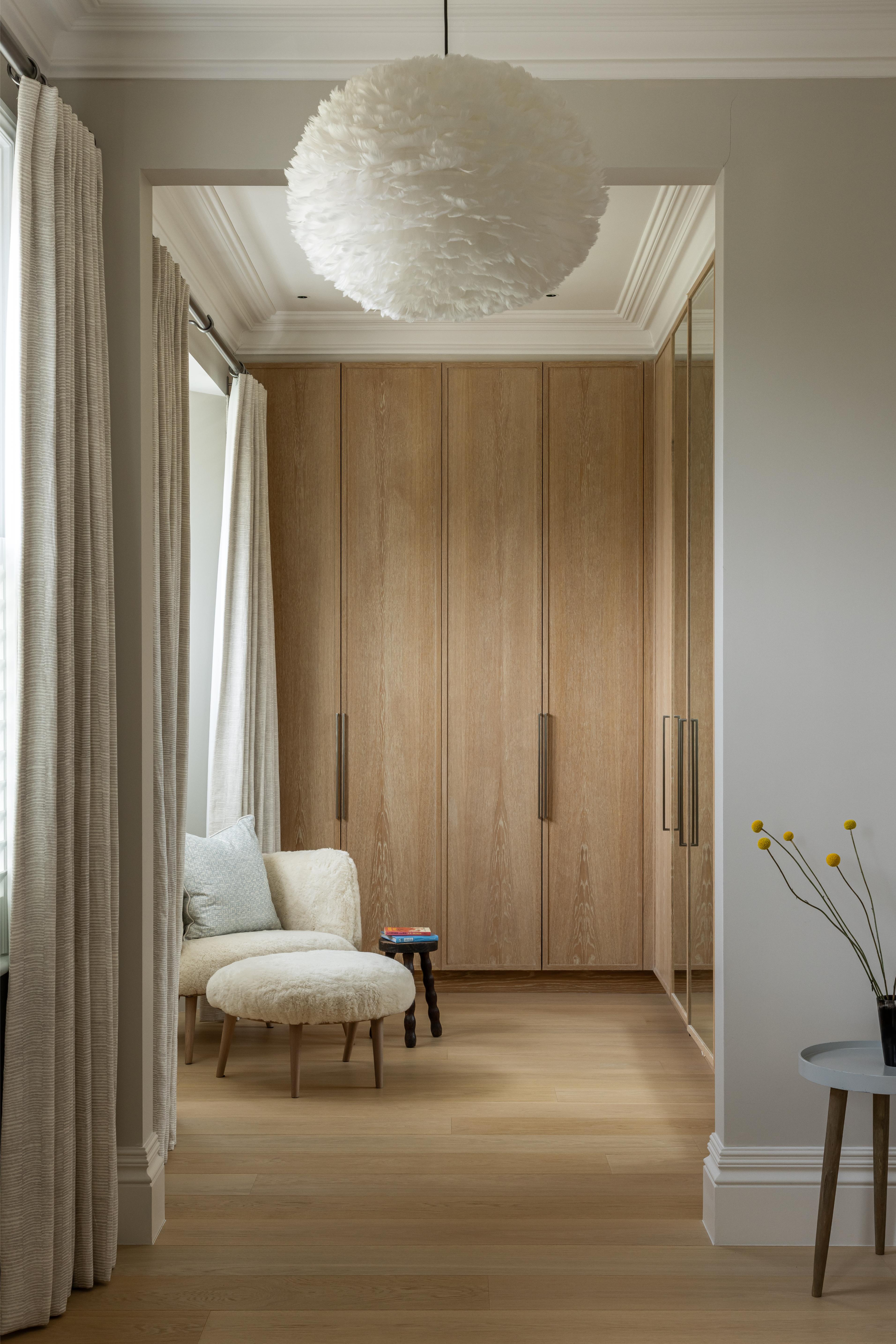 A wardrobe with timber doors in a bedroom with linen curtains, shearling chair and feather pendant light