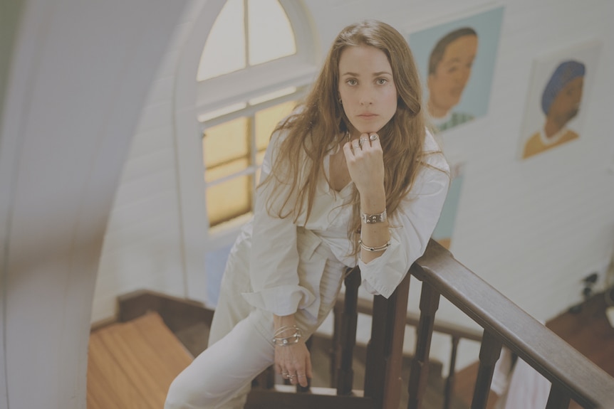 White woman in white shirt & pants with long light brown hair poses on a stairwell, one hand under chin.