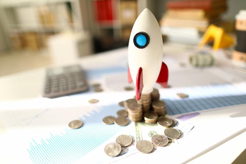 A toy rocket readying for launch atop messy stacks of coins and paperwork displaying financial data. 