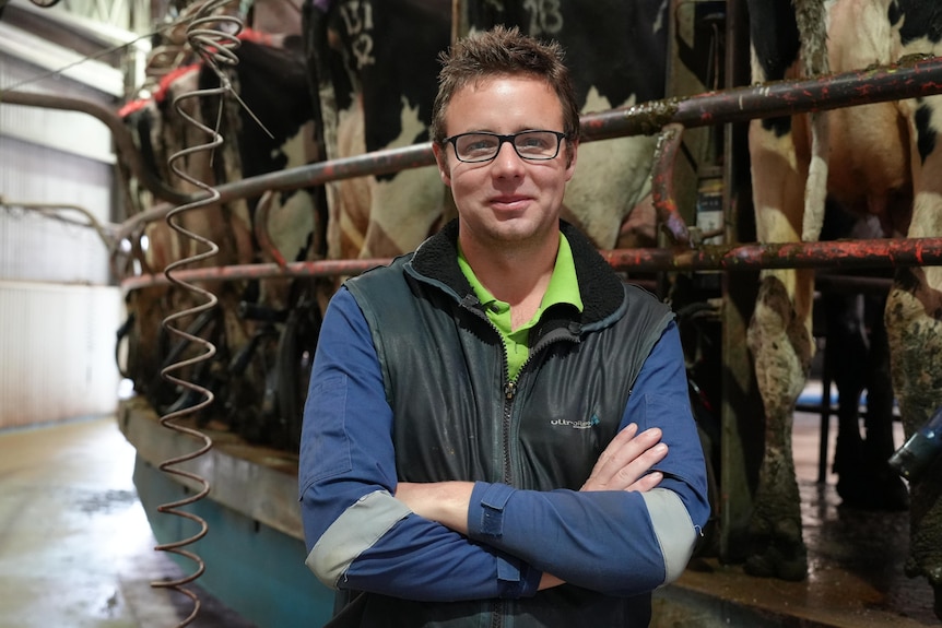 A dairy farmer stands with arms crossed with cows behind him in pens