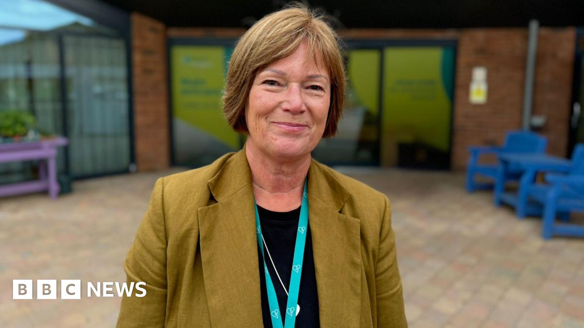 A woman with short brown hair stands on a paved area outside a hospice building. She is wearing a brown jacket and black roundnecked top with a pale blue lanyard visible. There is coloured furniture behind her.