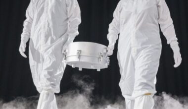 Lunar quarantine facility: Two men in white suits carrying a silver canister between them, in a dark area with white mist boiling up from the floor.