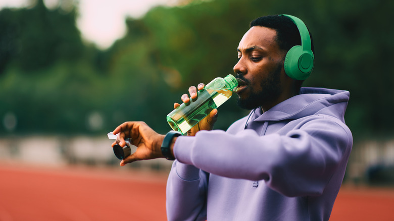 An individual in a light purple hoodie drinks from a green water bottle while they check their smartwatch on their other wrist, the individual appears to be on a running track