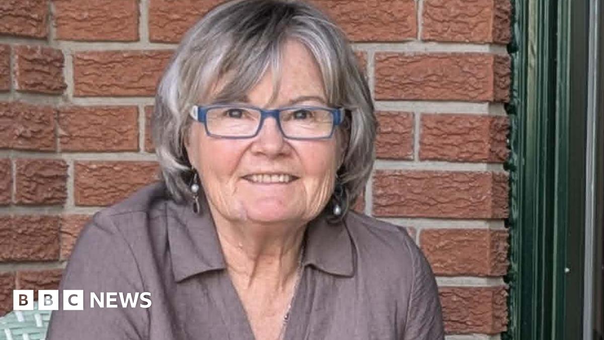 An image of Elizabeth Doran. She is smiling and has a short grey bob with bangs, blue-framed eyeglasses and dangling earrings. She is wearing a collared linen grey-mauve shirt, and behind her is a brown brick backdrop.