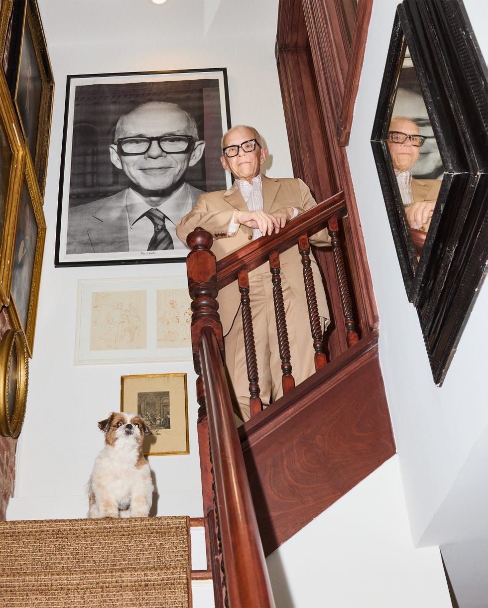 Man in glasses and tan suit stands on staircase with small dog below.