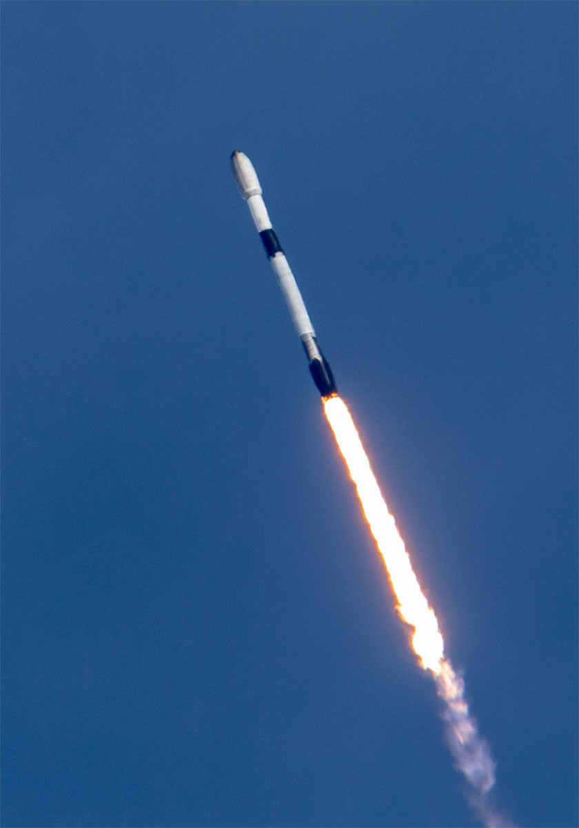 a white and black rocket set against a blue sky climbs toward space on a bright orange-white plume