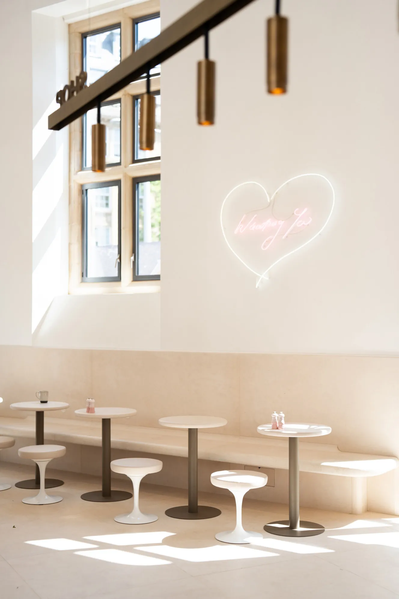 A cafe interior with light-colored tables and stools, a wall-mounted bench, and a pink neon heart sign.