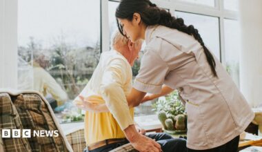 A young female carer helps an elderly male to get dressed by pulling on his jumper, as he sits in an armchair.