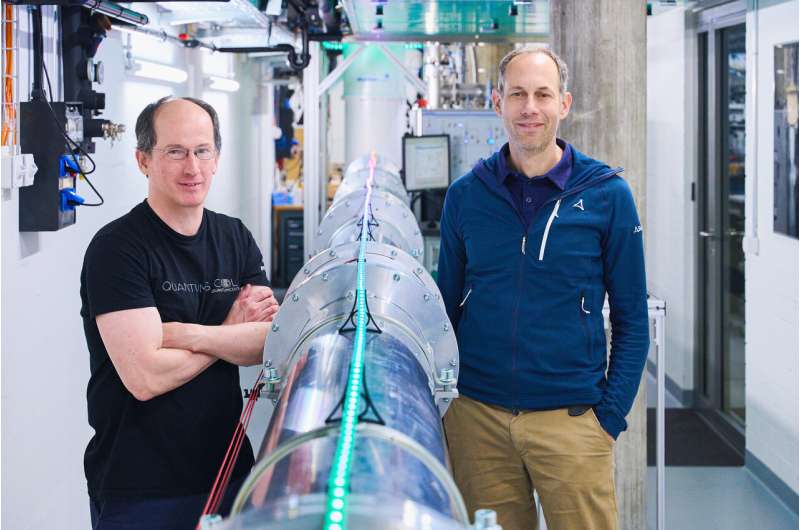 Two men stand next to a metal cylindrical tube in the middle of a laboratory