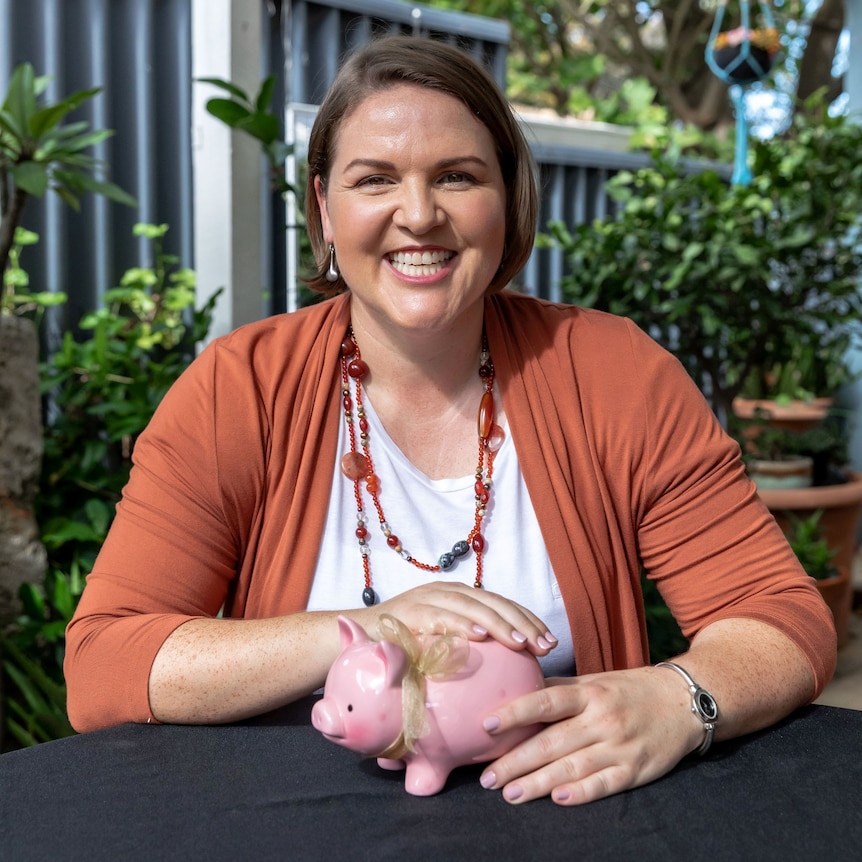 Lacey smiles as she sits at a table outside, holding a pink piggy bank.
