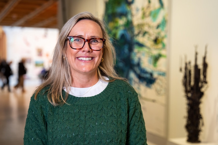 Rebecca Andrews, blonde hair, glasses, wearing a white shirt and green knit, smiling while standing in a gallery.