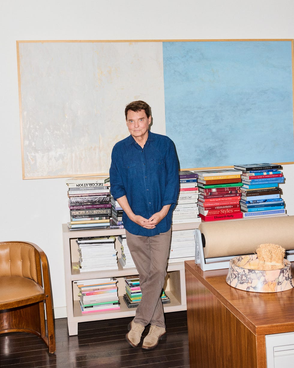 Person in a blue shirt standing in a room with stacks of books and abstract paintings on the wall.