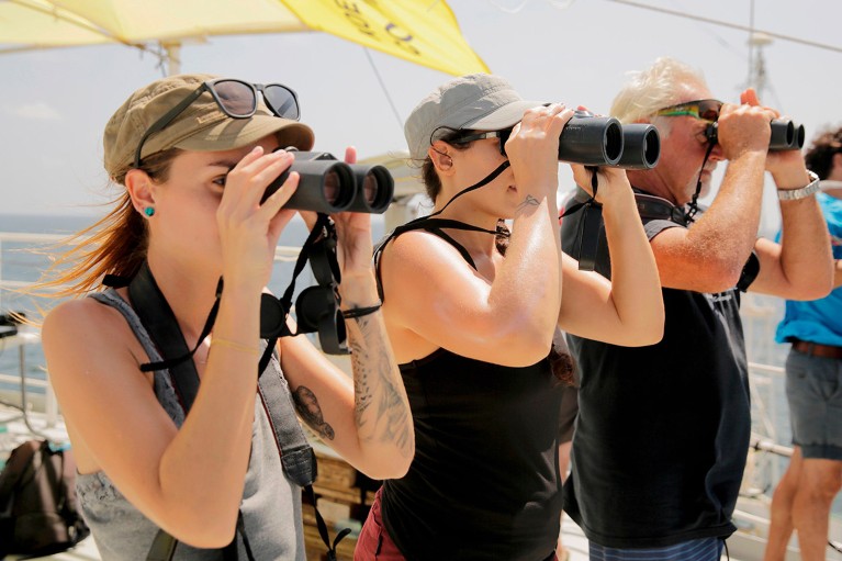 A line of three marine biologists, two women and one older man, use binoculars to look for whales from a boat off the French Guiana coast.
