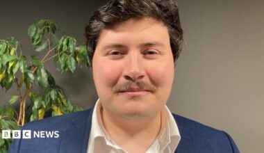 A close up of Deputy Tom Rylatt. He has short black hair, a brown moustache and is wearing a white shirt and navy jacket.