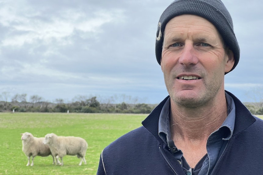 A man wearing a beanie in a green paddock with sheep in the background