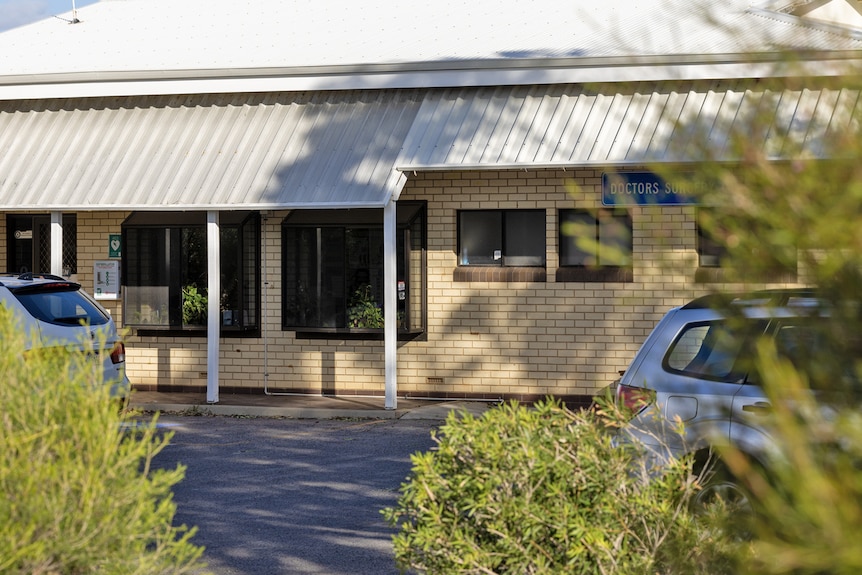 A brick building with two cars out the front parked up behind bushland.