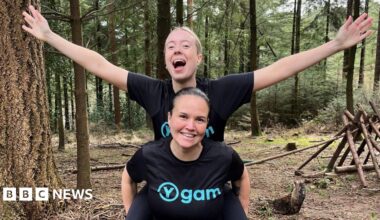 Sisters Michelle Kelly and Emma dressed in running clothing. They are in a forest, with one carrying the other on their back and arms stretched wide. Both are smiling at the camera.