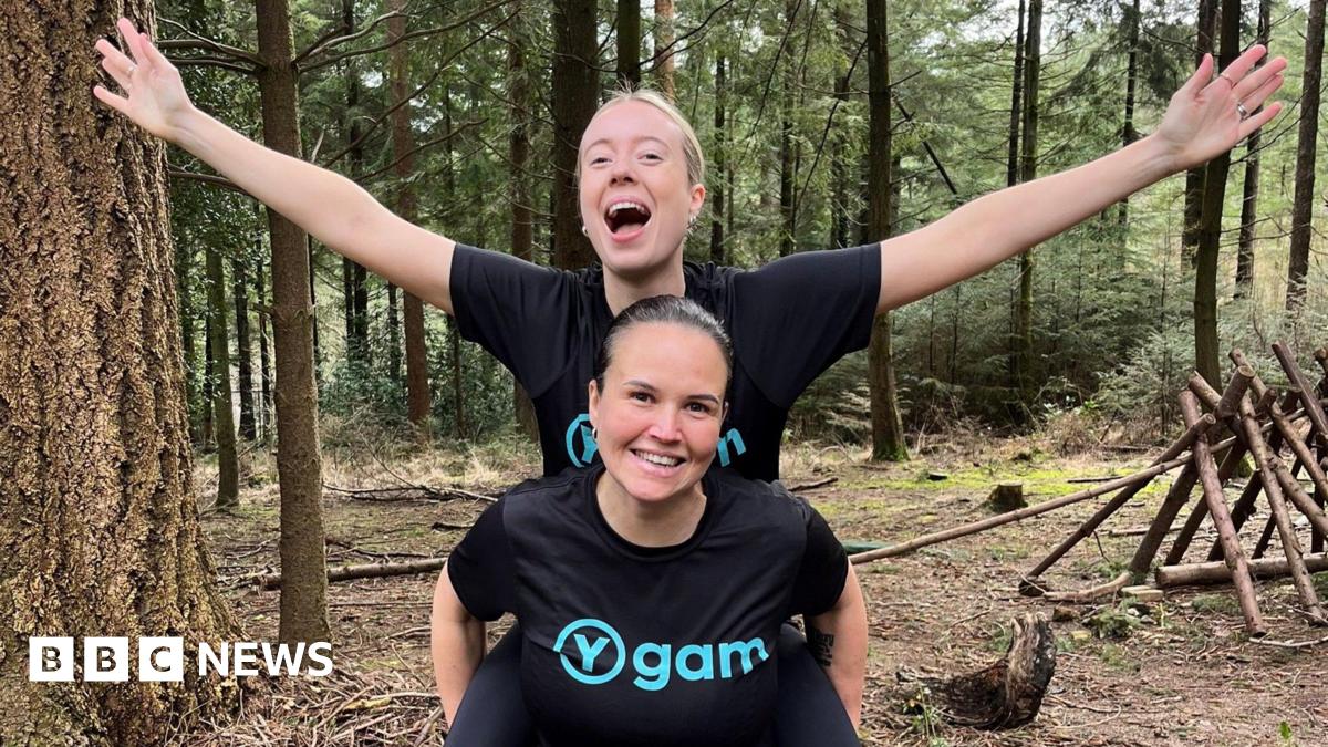 Sisters Michelle Kelly and Emma dressed in running clothing. They are in a forest, with one carrying the other on their back and arms stretched wide. Both are smiling at the camera.