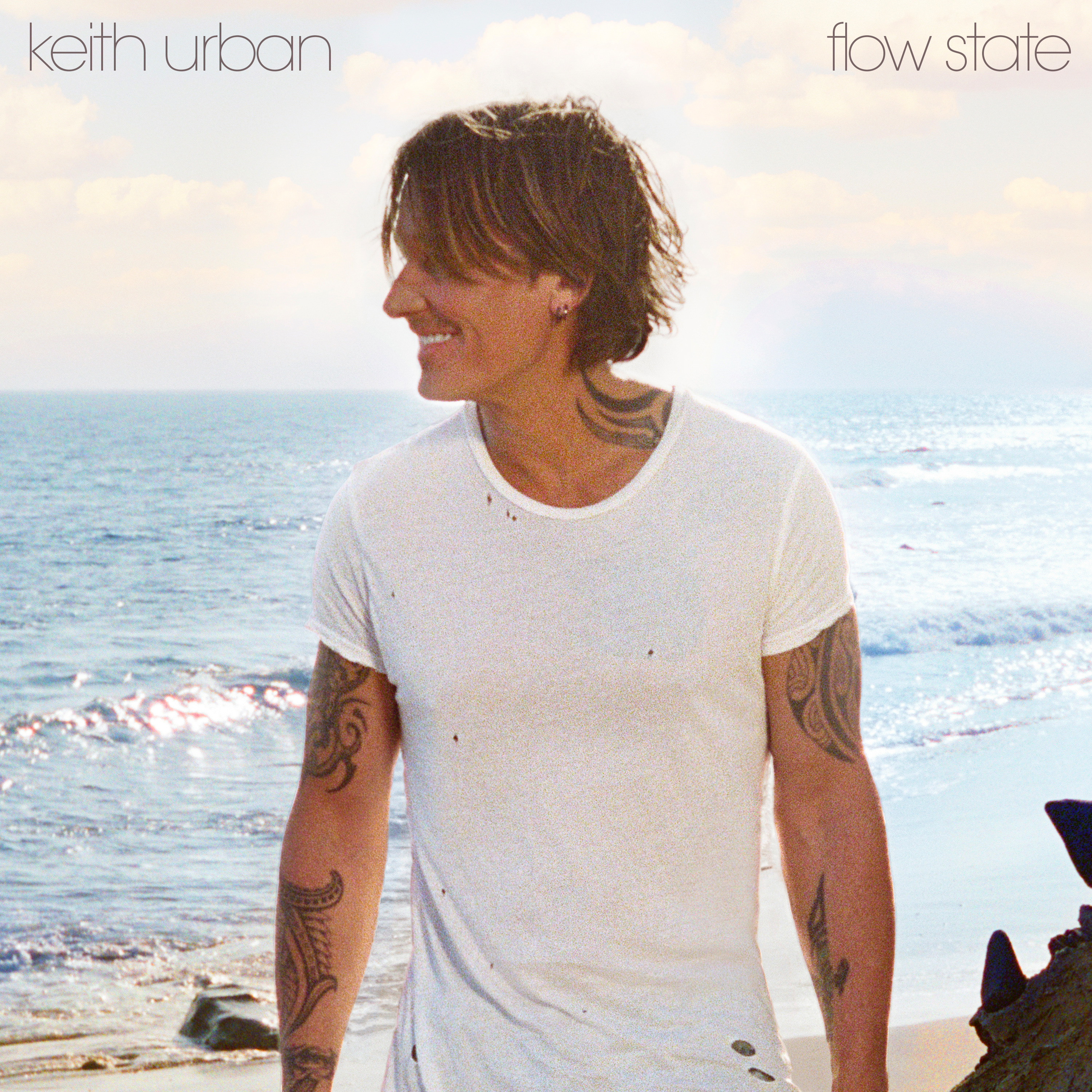 Album cover of a tattooed Keith Urban in white T-shirt standing by the ocean, text reads “keith urban – flow state”