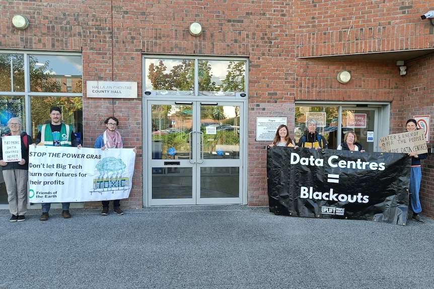 A small cluster of people outside a building hold banners opposing data centres.
