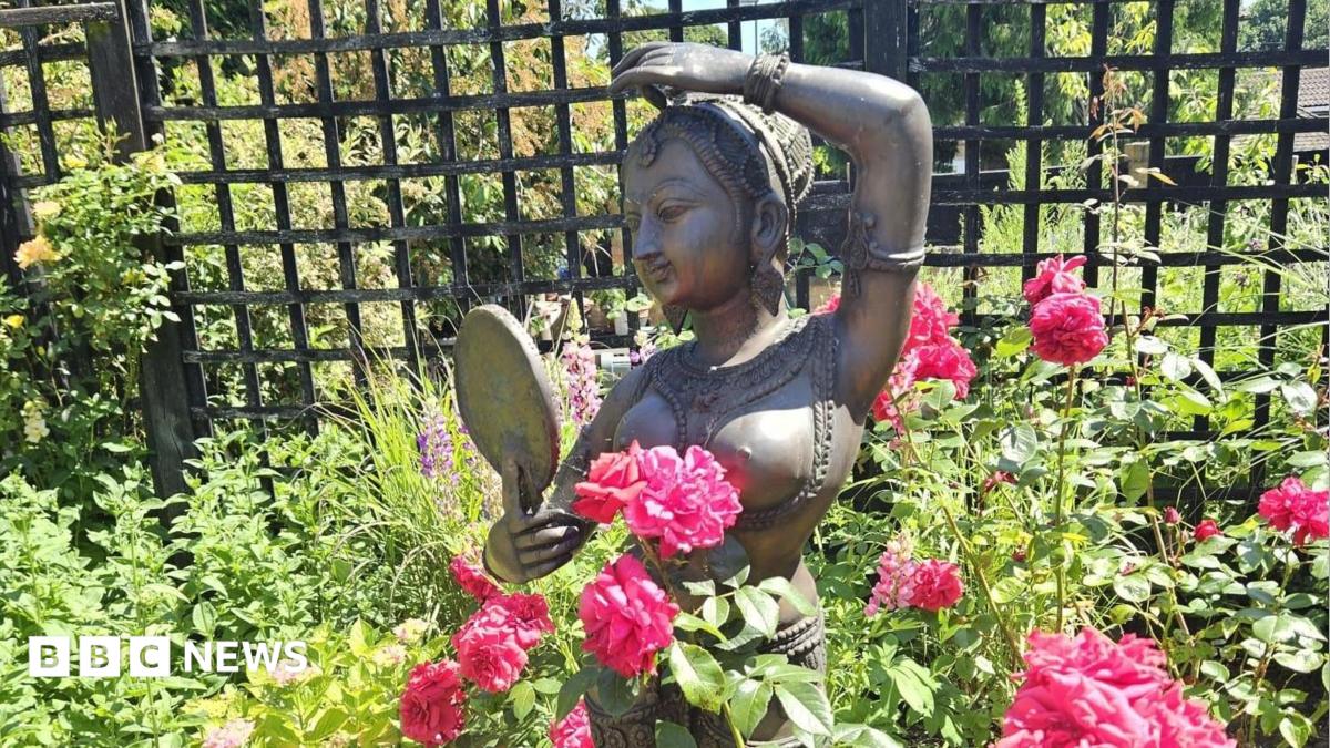 A bronze statue of an Indian woman is placed among pink flowers in a garden. She is designed as if wearing ornate clothing and looking at herself in a mirror. There are shrubs behind her and a trellis fence.