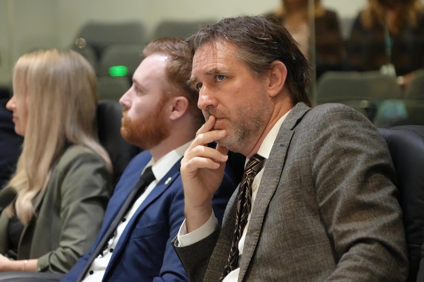 A man in a suit sitting with other people with a serious facial expression and leaning on his elbow with face resting in hand.