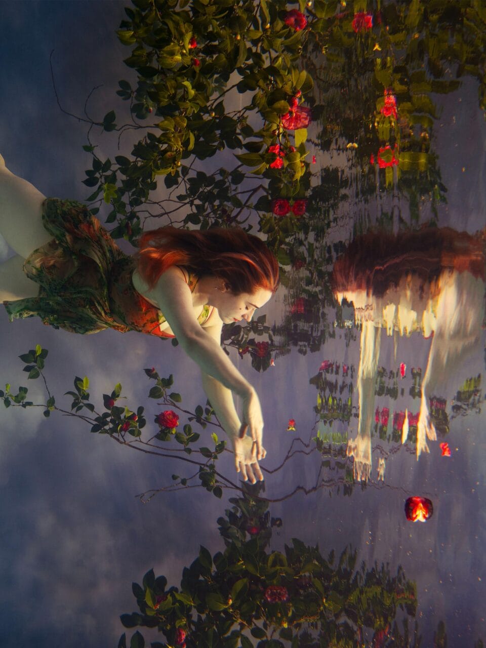 a photograph by Tamara Dean of a woman with red hair drifting in water, among branches with leaves and pink flowers