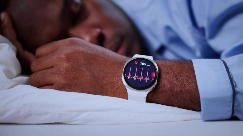 A man sleeps on a bed while his smartwatch shows his heart rate.