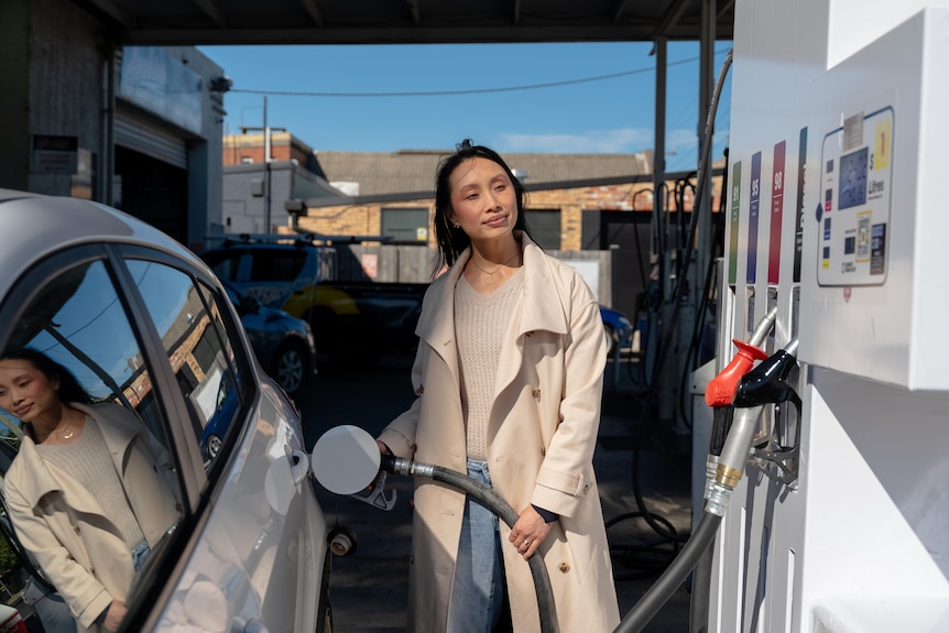 A woman stands wearing a beige top and trench coat at a fuel pump. She's filling up her silver hatchback car.