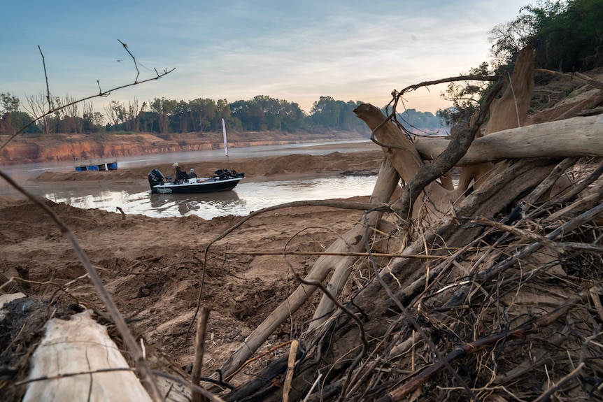 Foreground are broken tree branches, piles of mud, with a small fishing black fishing boat with two people in the background.
