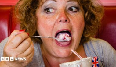 One of Emma's photographs. She pictures a woman with flowy ginger hair eating an ice cream with a long spoon. She is putting the ice cream in her mouth - whilst sitting on a red leather seat. A Union Jack flag sits limply out the top of her ice cream.