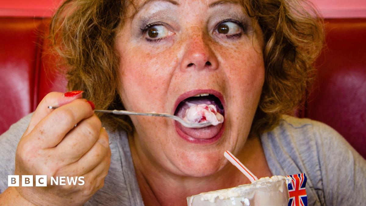 One of Emma's photographs. She pictures a woman with flowy ginger hair eating an ice cream with a long spoon. She is putting the ice cream in her mouth - whilst sitting on a red leather seat. A Union Jack flag sits limply out the top of her ice cream.