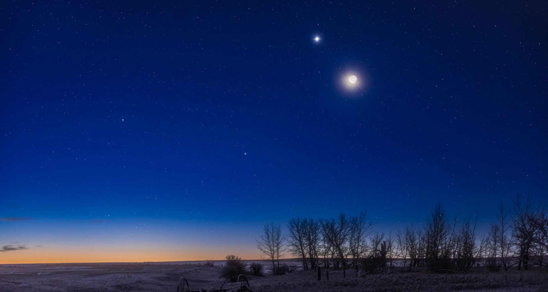 The moon shines in the evening sky with a bright star-like planet glowing close to its upper left above a snowy field lined with trees. The glow of the setting sun can be seen brightening the horizon.